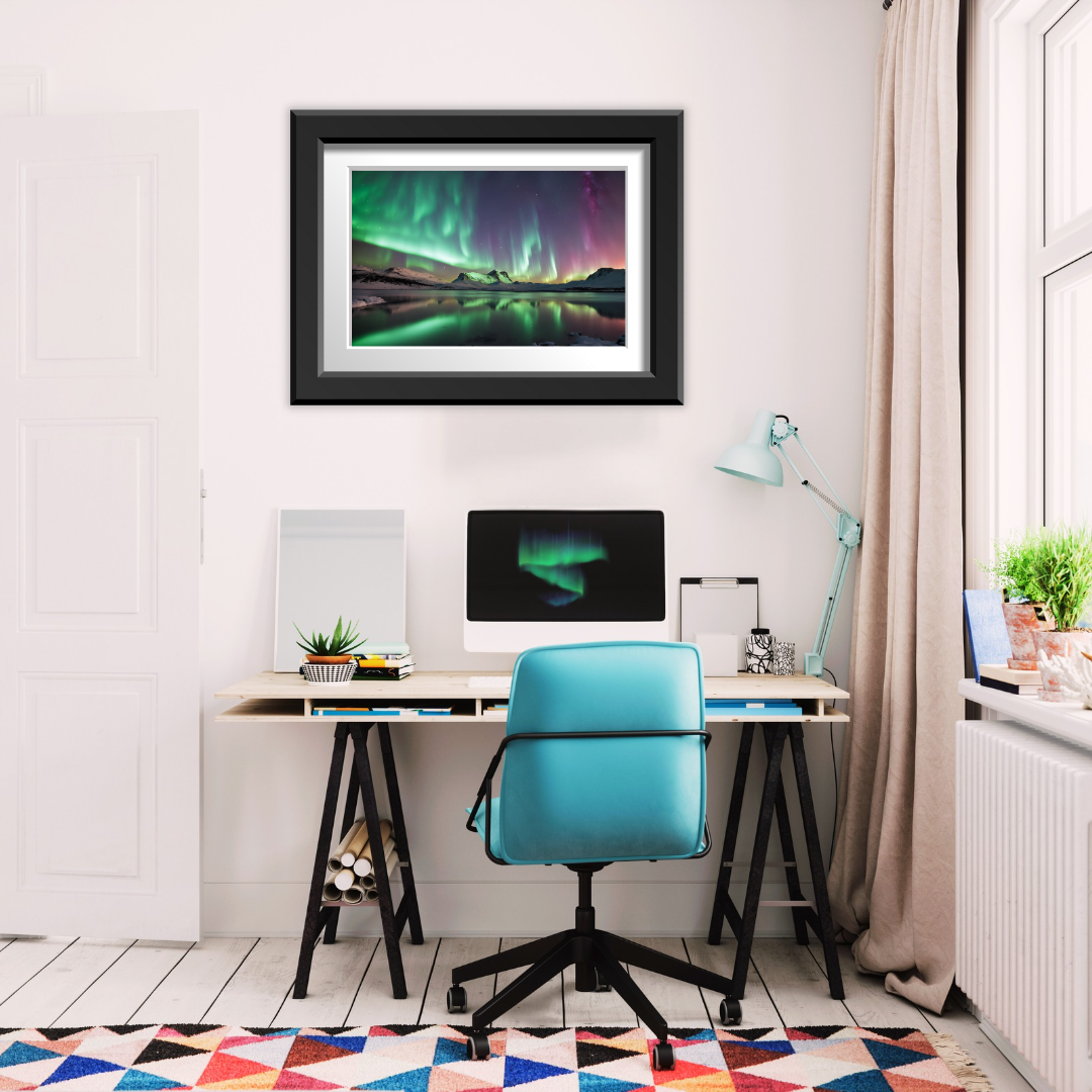 Framed Aurora Borealis print above a minimalist desk with lamp, plant, and computer in a home study setting.