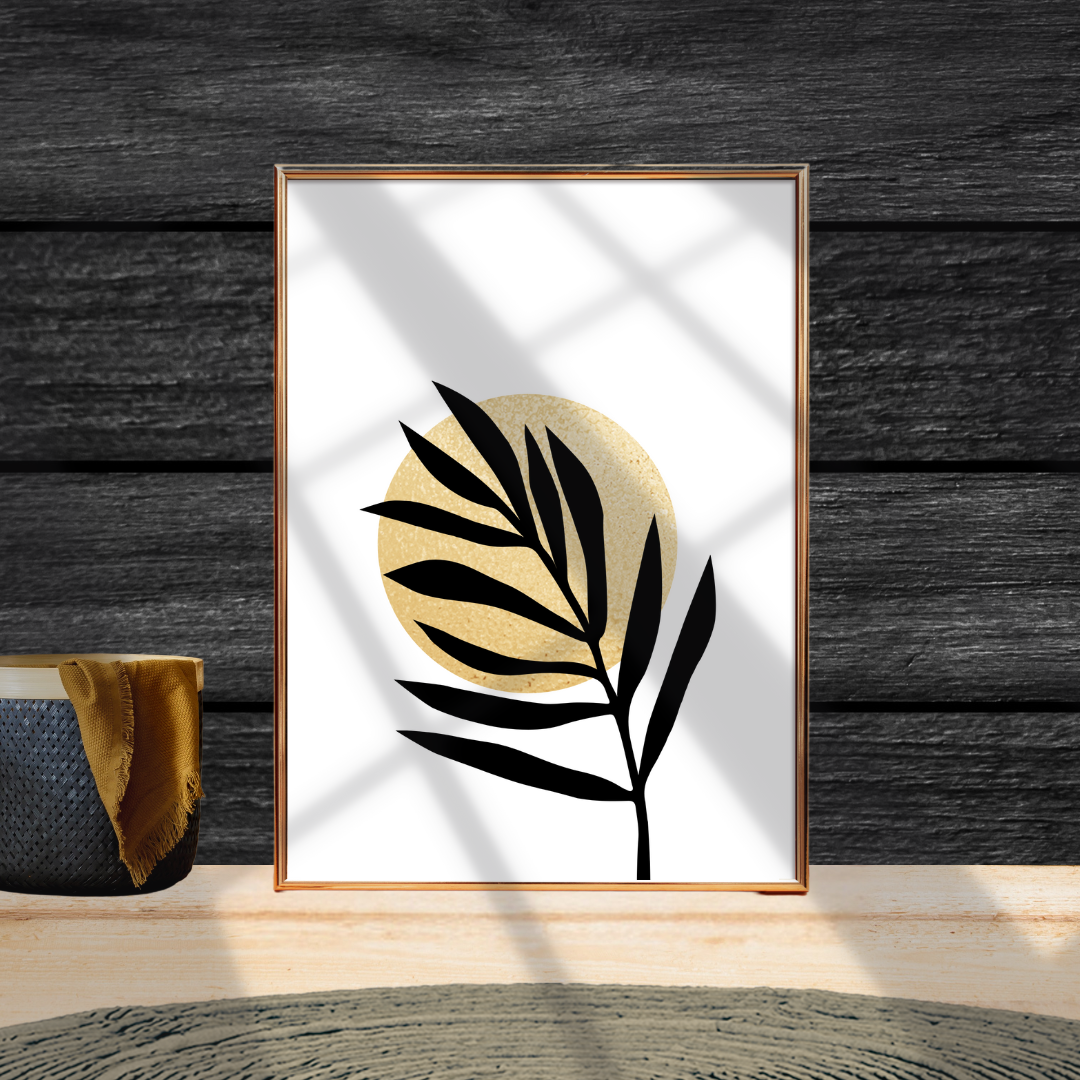 Framed botanical artwork with black leaf design with a gold sun on a white background against a dark wooden wall.