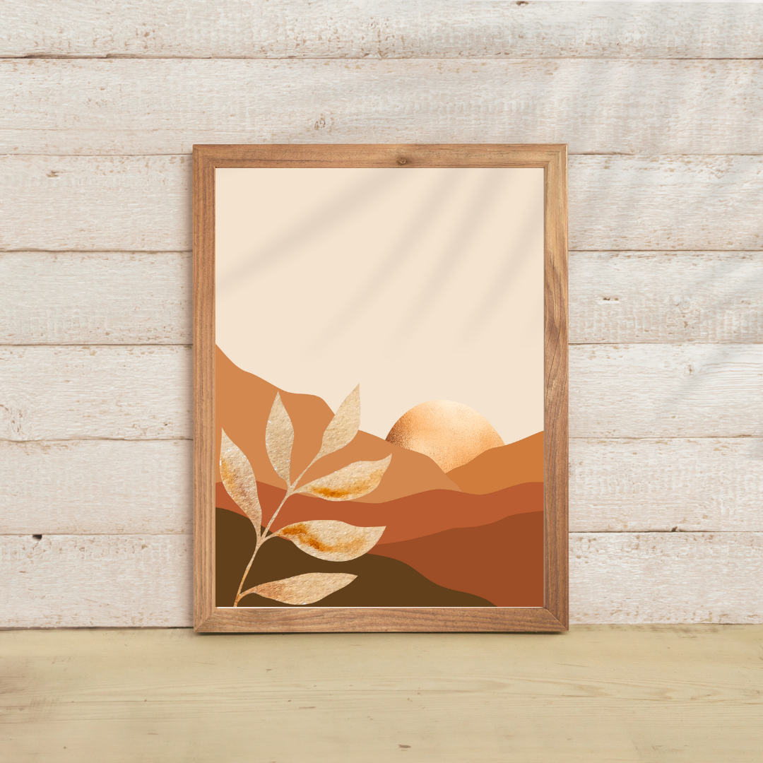 Close-up of a layered mountain wall art print in earth tones featuring a golden sun in a wooden frame resting on a light wood backdrop.