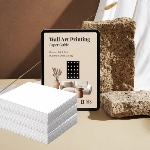 Stack of white paper sheets with a tablet displaying a 'Wall Art Printing Paper Guide' in a minimalistic setting.