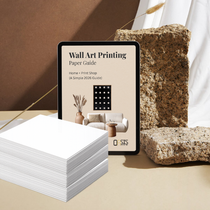 Stack of white paper sheets with a tablet displaying a 'Wall Art Printing Paper Guide' in a minimalistic setting.